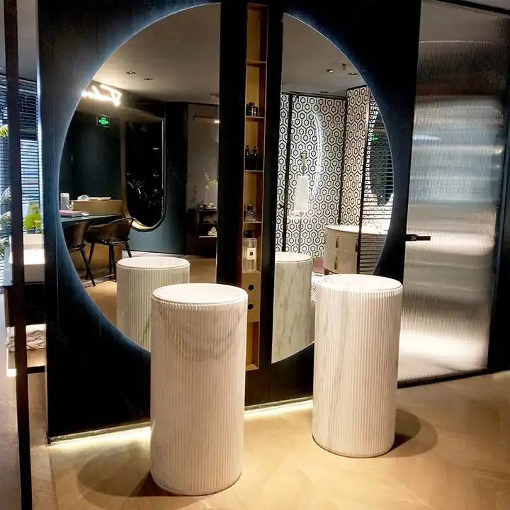 Elegance Luxury Half Moon Glow mirrors with backlighting flanking a tall shelf behind fluted marble pedestal sinks in a showroom.
