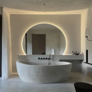 A Luxury Organic Circle Backlit 4/6 Moon Wall Mirror glowing behind a modern stone bathtub in a minimalist bathroom.