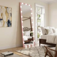 Tall pink-framed mirror with round lights leaning in a bright bedroom, reflecting a white bed and balcony door.