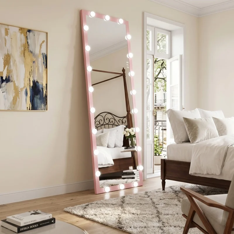 Tall pink-framed mirror with round lights leaning in a bright bedroom, reflecting a white bed and balcony door.