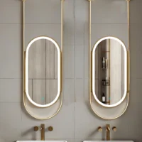 Double oval backlit mirrors with gold frames mounted on a light gray tiled bathroom wall, reflecting built-in shelves and toiletries above two gold faucets and sinks.