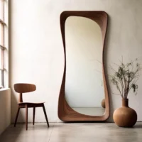 A Japandi Wood Organic Floor Mirror with a thick curved frame, leaning against a white wall next to a wooden chair.