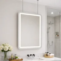 Rectangular backlit bathroom mirror above a marble vanity with a sink, flowers, and a glass-enclosed shower to the right.