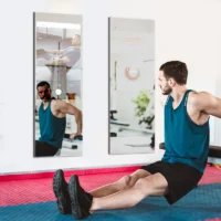 Man in a blue tank top doing sit-ups on a mat in a gym, facing wall mirrors reflecting his silhouette.