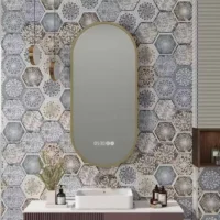 Oval mirror with a gold frame above a white rectangular sink on a striped vanity, set against a wall of blue-gray hexagonal patterned tiles and decorative pendant lights to the left.
