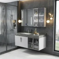 Modern bathroom with a gray marble countertop vanity, wall cabinet with glass doors, and open shelves beneath a dark tile wall. A glass shower to the left and a large window to the right frame the scene, with gold pendant lights overhead.