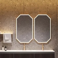 Two octagonal mirrors with gold rims above a white countertop sink in a modern stone bathroom. A towel rack and bottles sit to the left.