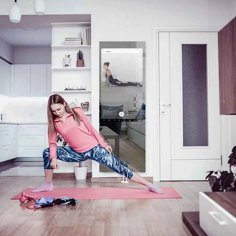 Woman in a pink top and blue patterned leggings performs a side lunge on a pink yoga mat in a bright home space, with a yoga video playing on a wall-mounted screen/mirror.