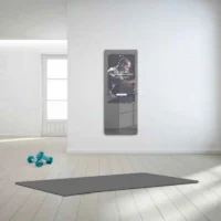 Bright minimalist room with a wall-mounted smart mirror showing a workout poster, blue dumbbells, and a large gray mat on the floor.