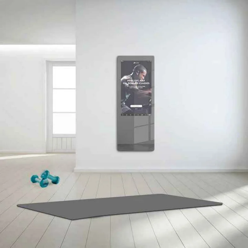 Bright minimalist room with a wall-mounted smart mirror showing a workout poster, blue dumbbells, and a large gray mat on the floor.